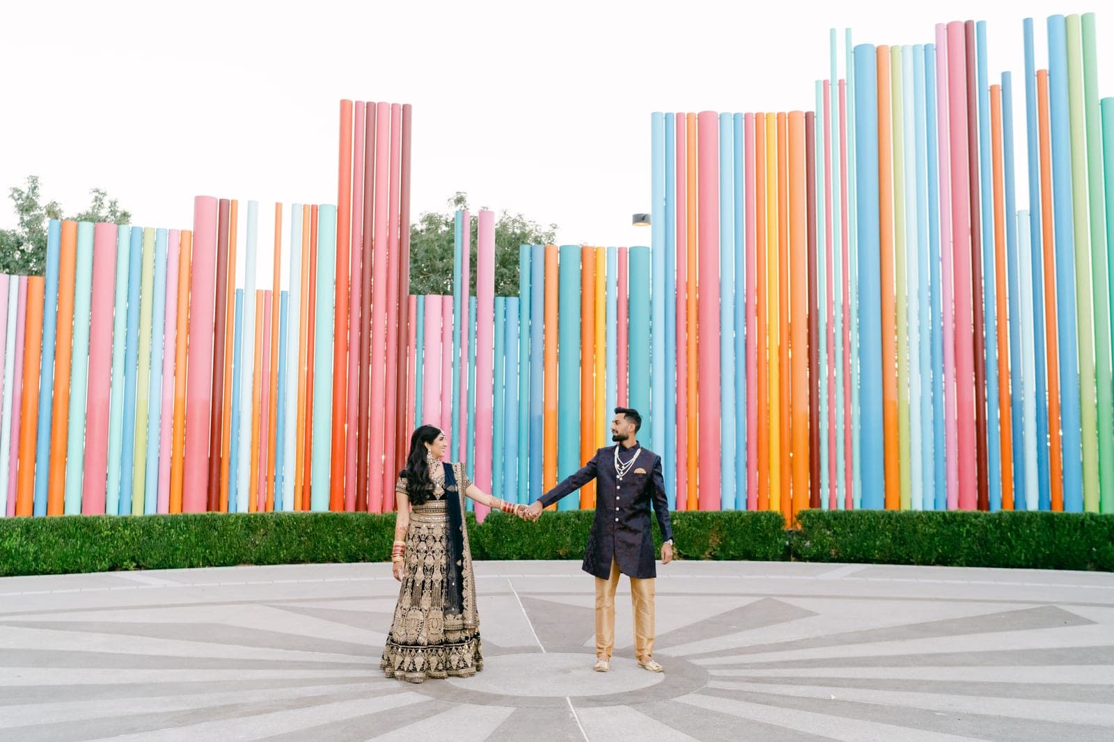 A couple in South Asian wedding attire hand in hand before Tim Bavington's repeating color columns at The Smith Center's Symphony Park, photographed by Amber and Co Photo